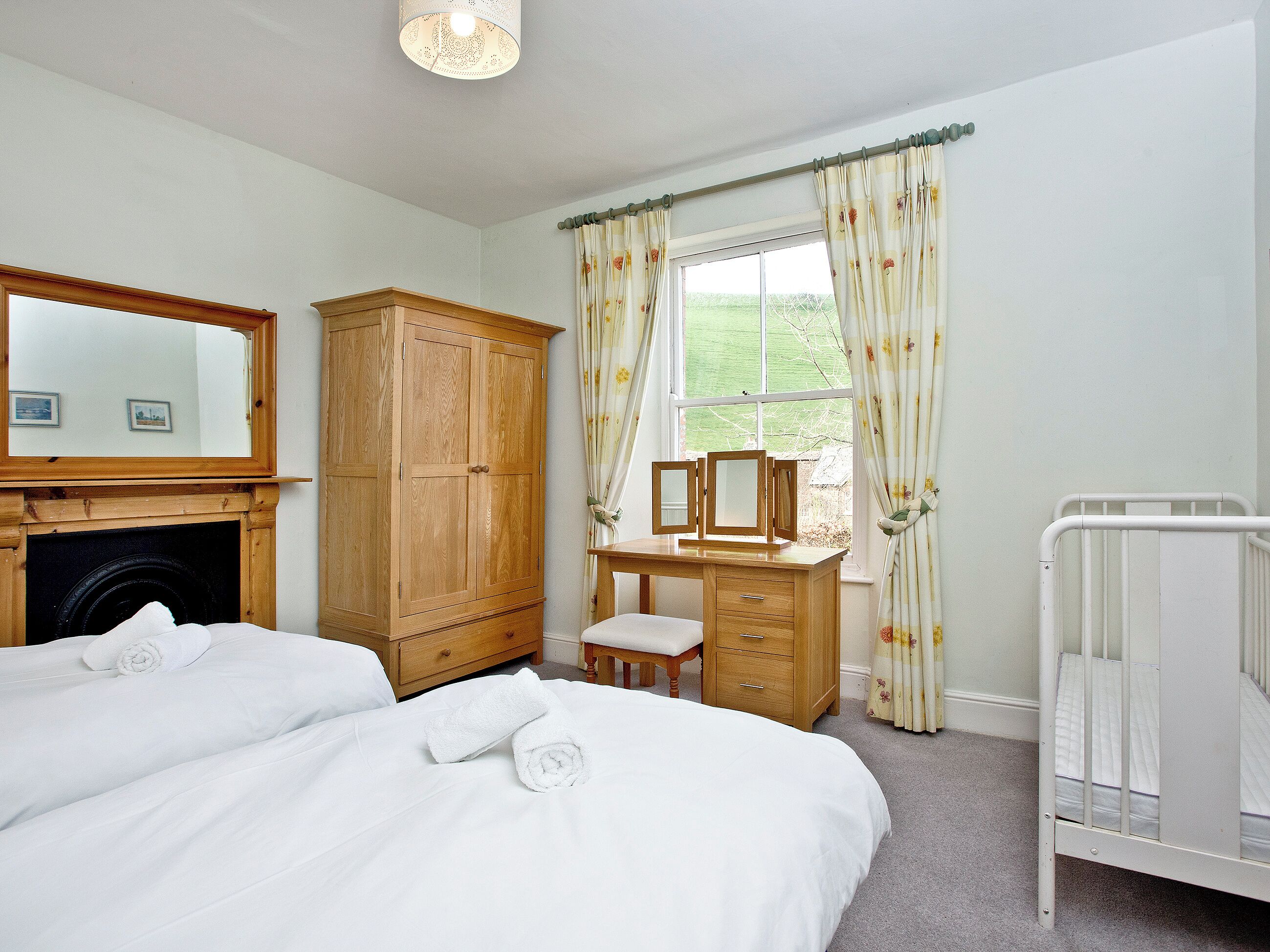 A bedroom with a bed, a wooden wardrobe, a desk with a chair, a crib, and a window with curtains.