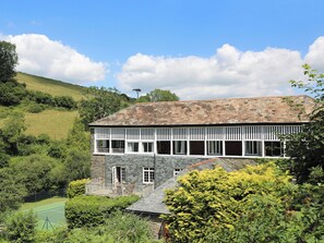 A two-story stone building with a balcony, surrounded by lush greenery and a tennis court. - Tuckenhay, Mill House - FUB (Bow Creek, between Dartmouth and Totnes)