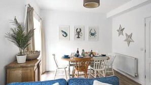 Une pièce lumineuse et bien éclairée, avec une table à manger dressée pour quatre personnes, entourée de chaises blanches. Trois tableaux encadrés ornent le mur, une plante en pot est posée sur un meuble en bois, et un luminaire est fixé au plafond.
