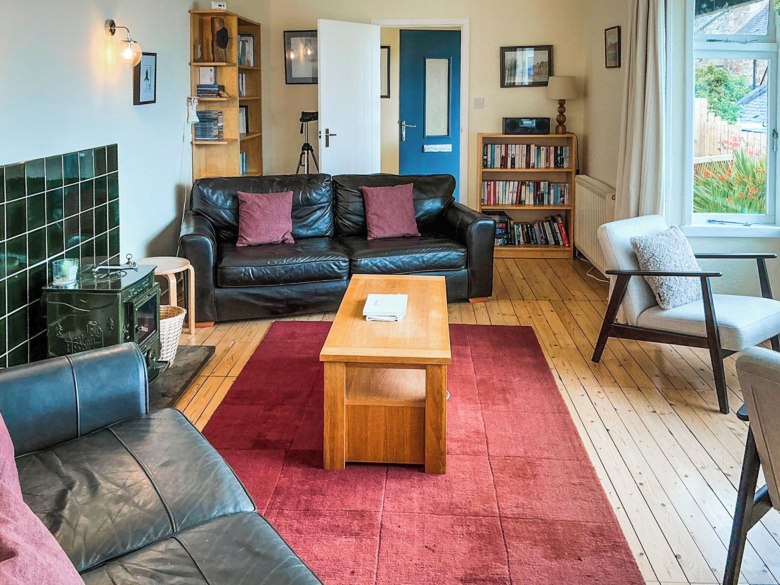 A cozy living room with a black leather sofa, a wooden coffee table, and a red rug. There are bookshelves filled with books and a blue door in the background.
