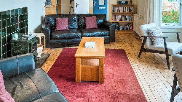 Een gezellige woonkamer met een zwarte leren bank, een houten salontafel en een rood vloerkleed. Er staan boekenplanken vol met boeken en in de achtergrond een blauwe deur.