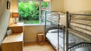 Een slaapkamer met een stapelbed, een bureau en een kruk, een raam met uitzicht op groen, en een lamp op het bureau.