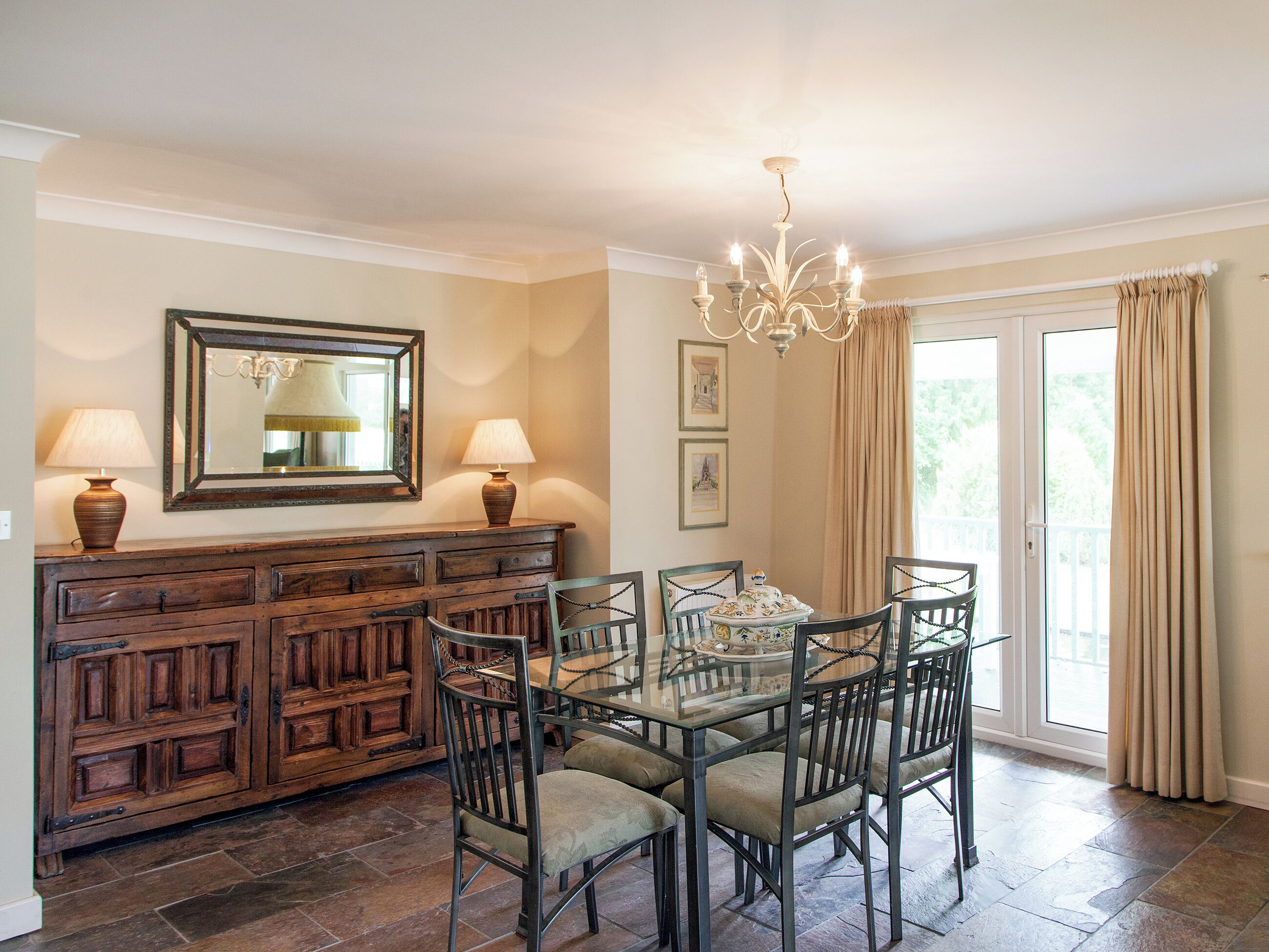 Un coin repas comprenant une table en verre, des chaises en métal, un buffet en bois et un lustre.