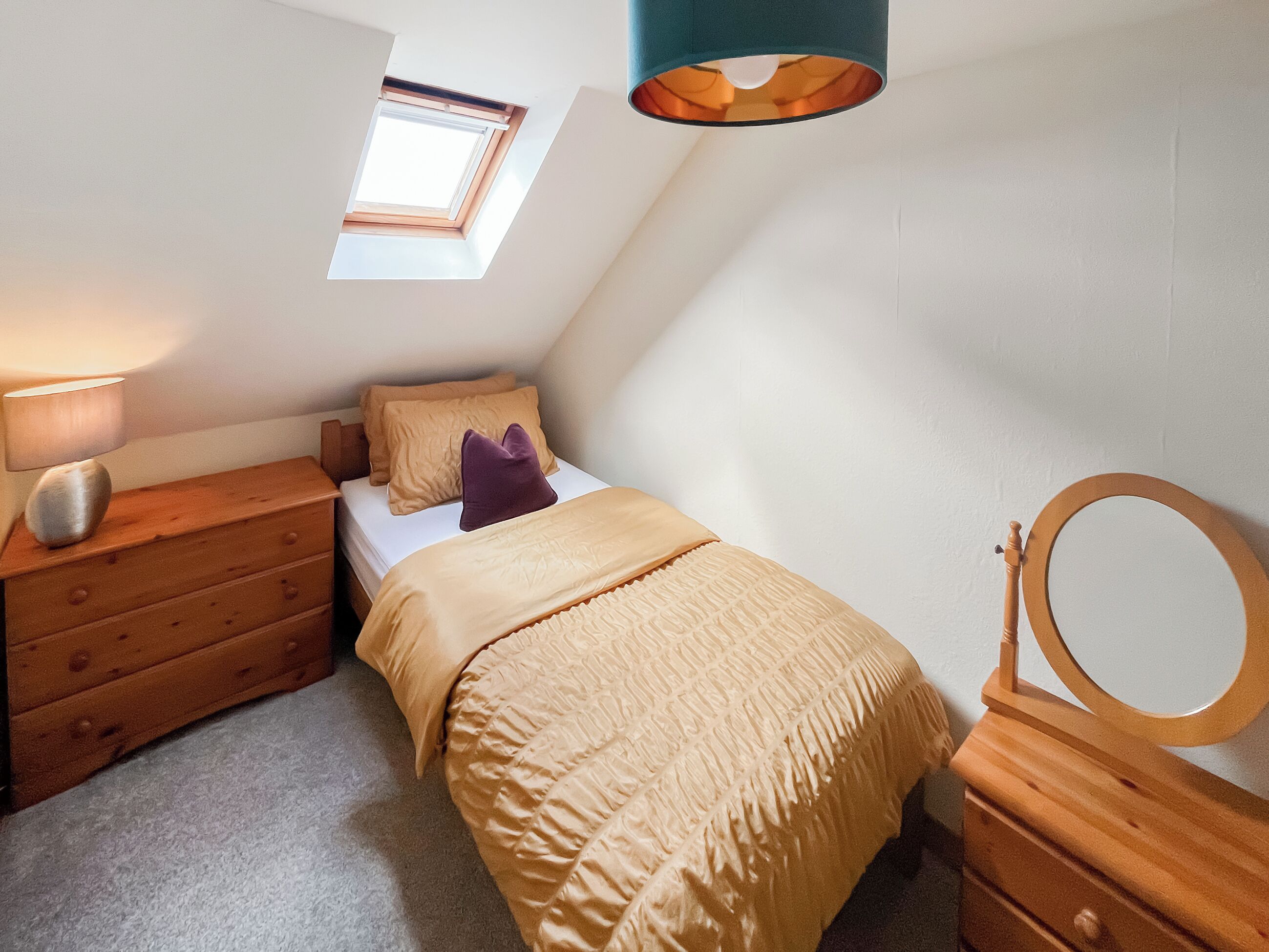 A bedroom with a skylight, a bed with a beige duvet, a wooden dresser, and a bedside table with a lamp.