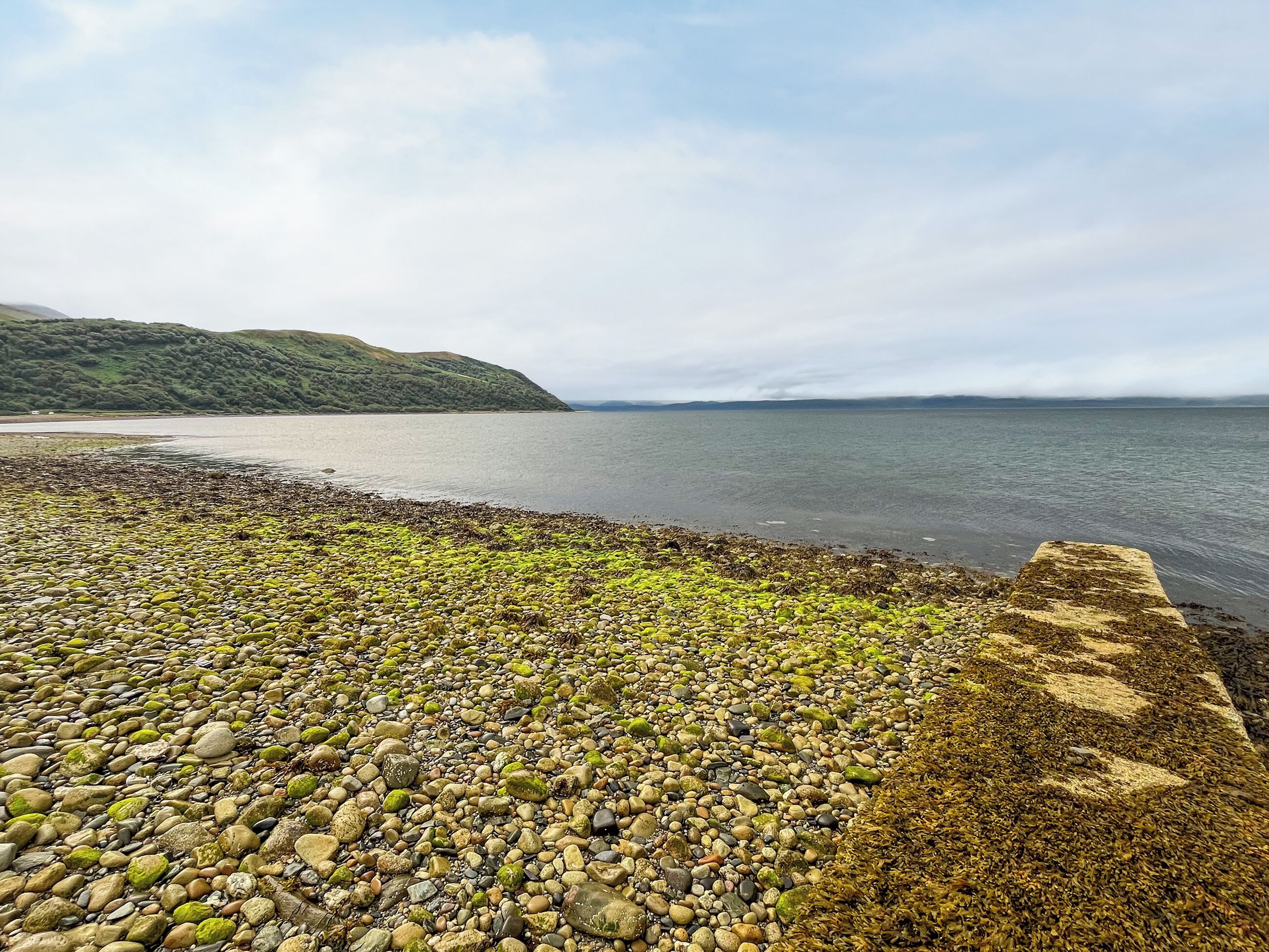 A pebbly beach with a stone wall and calm water.