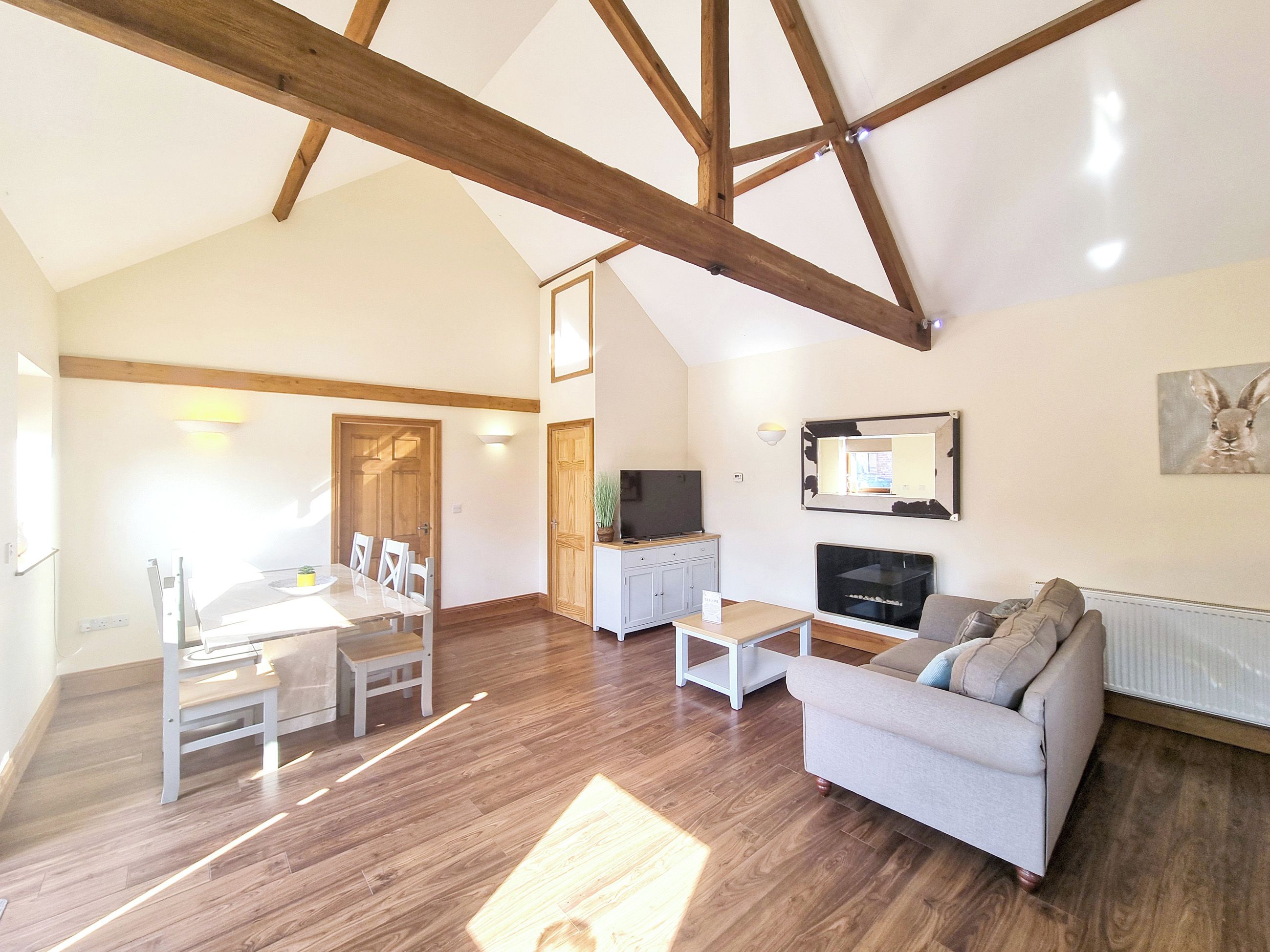 A spacious living area with a high ceiling, wooden beams, a dining table with chairs, a sofa, a television, and a fireplace.