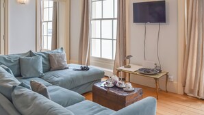 En stue med en lyseblå sofa, et lille bord med et tepotteskjul og et fjernsyn ophængt på væggen.