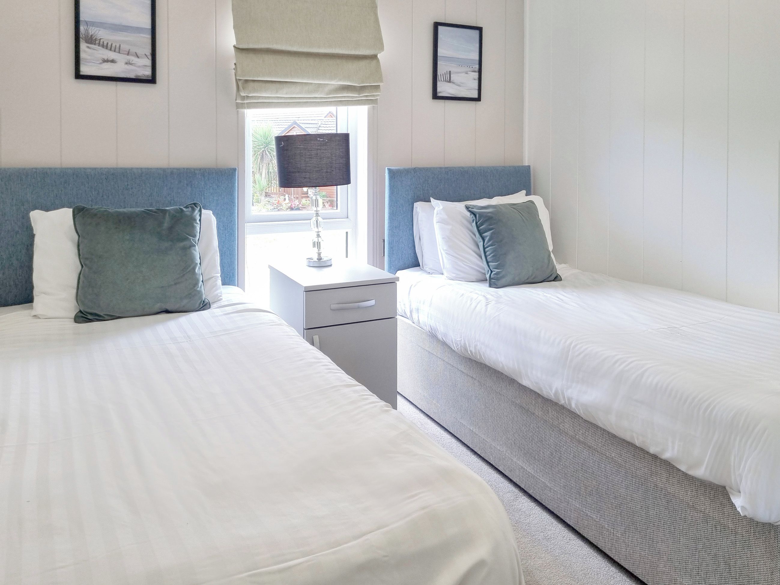 A hotel room with two beds, a nightstand, a lamp, and a window with a view of the outside.