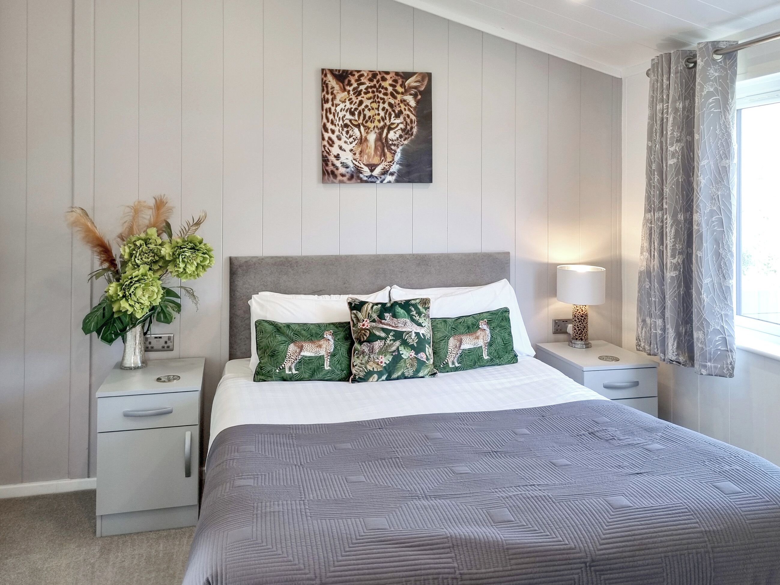 Une chambre à coucher bien rangée, avec un grand lit, deux oreillers à motifs animaliers, un tableau représentant un léopard accroché au mur, une table de chevet avec une lampe et une fenêtre avec des rideaux.