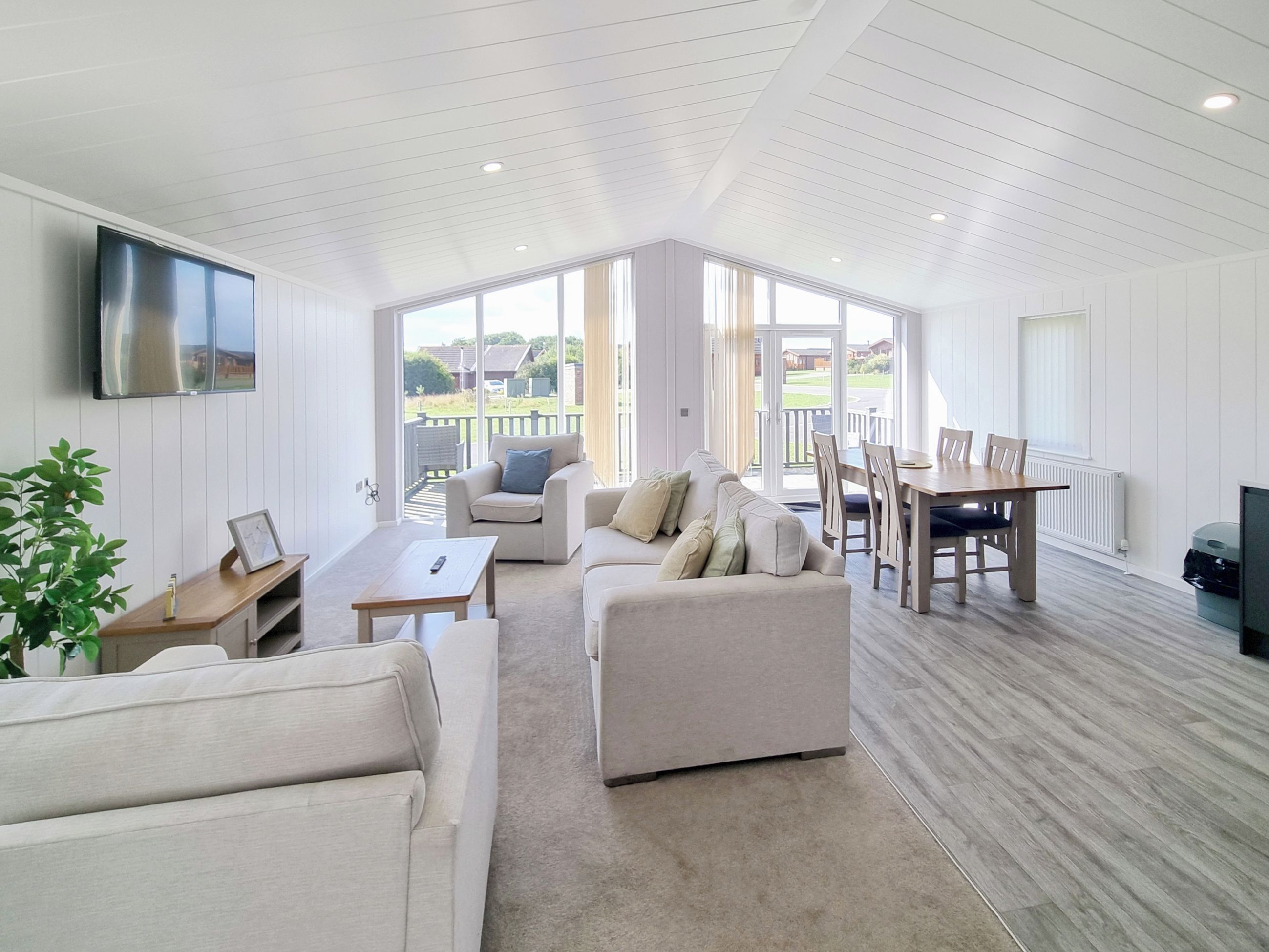 A spacious living area with a high ceiling, a flat-screen TV mounted on the wall, a sofa, a coffee table, and a dining table with chairs.