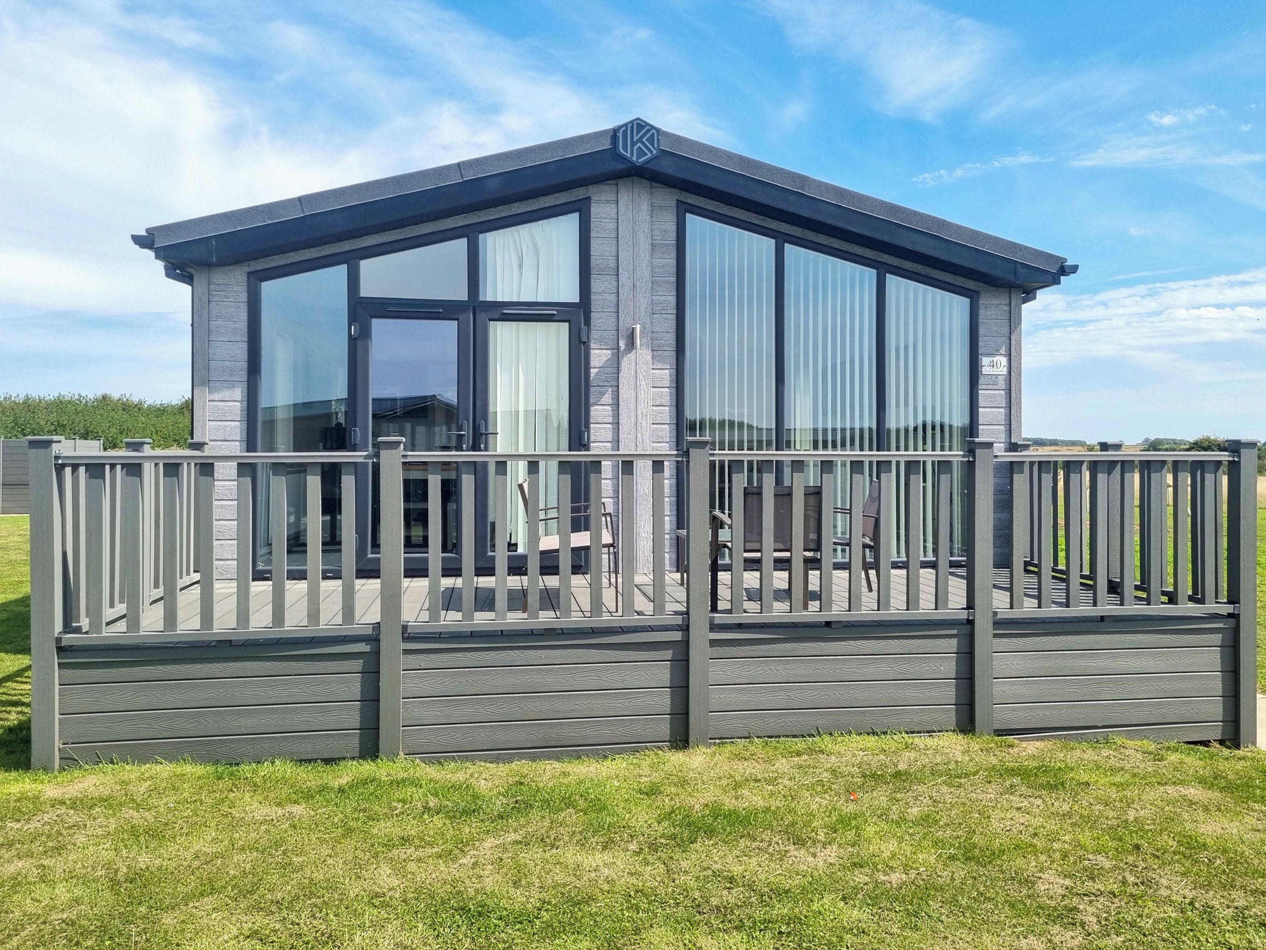 A modern, grey wooden cabin with large windows and a balcony, surrounded by a wooden fence.