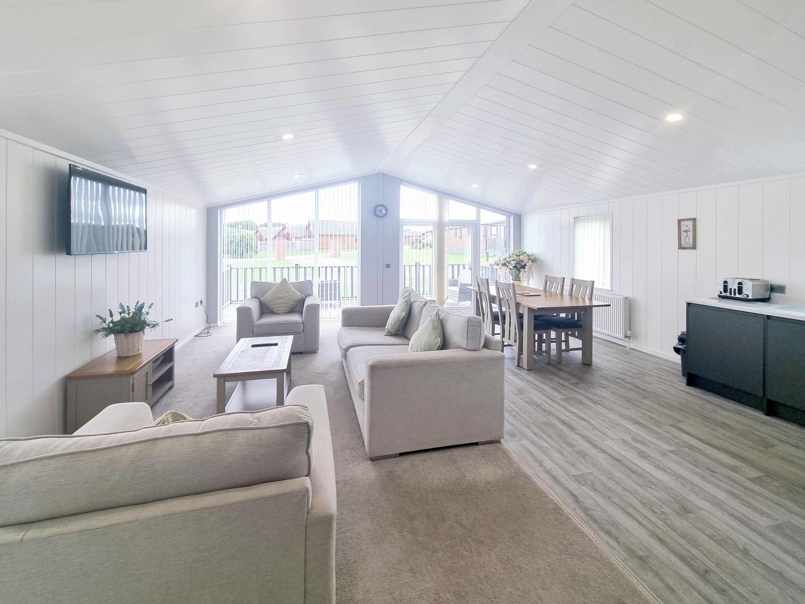 A spacious living area with a high ceiling, a dining table with chairs, a sofa, and a television mounted on the wall.