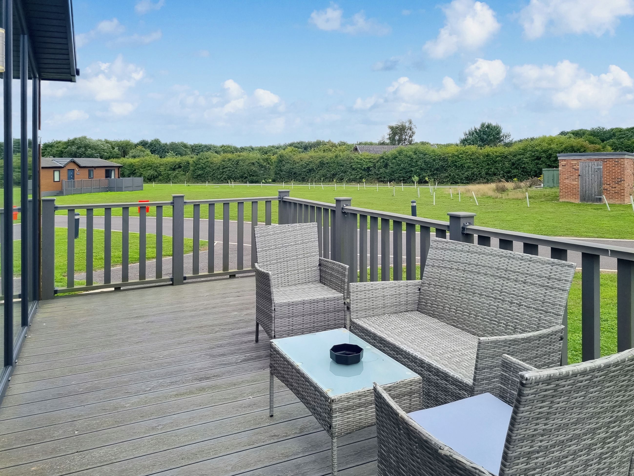 A deck with wicker furniture and a table, overlooking a green field and a brick building.