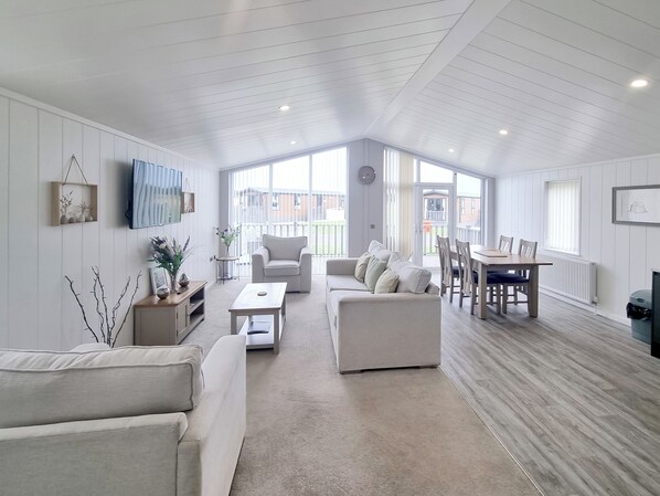 A spacious living room with a high ceiling, a dining area with a wooden table and chairs, and large windows allowing natural light.