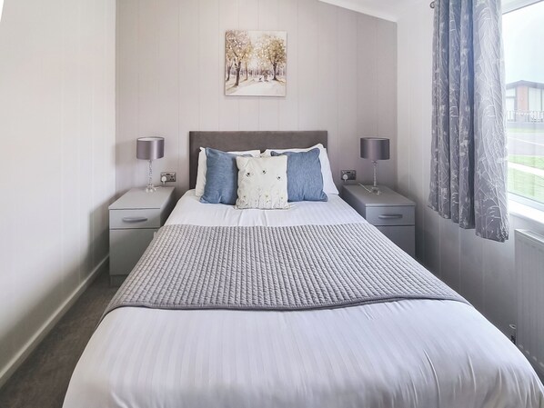 Un lit bien fait, recouvert d’une couverture matelassée, deux tables de chevet avec des lampes, un tableau accroché au mur et une fenêtre avec des rideaux.