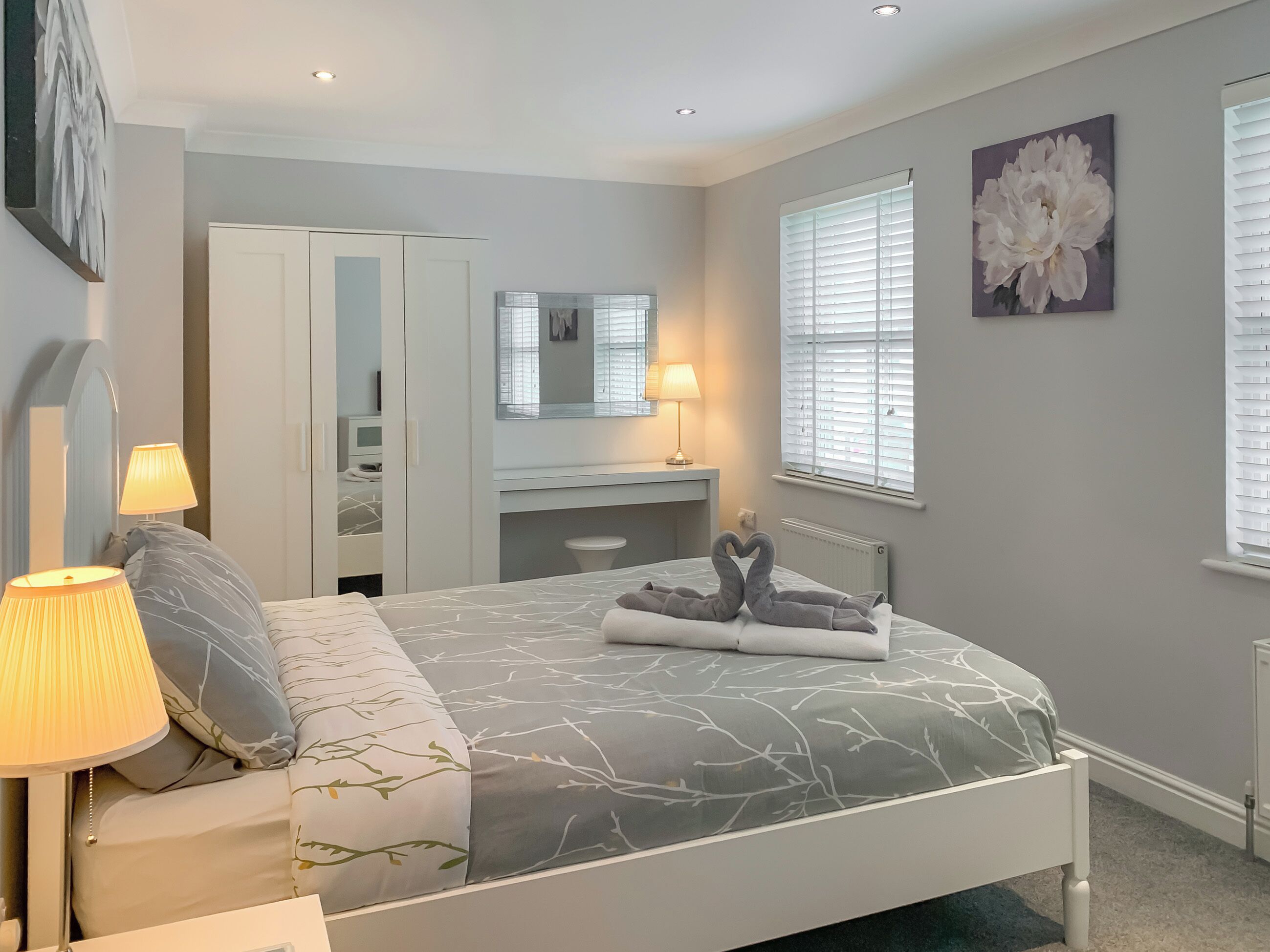 Een slaapkamer met een bed, nachtlampen, een dressoir, een spiegel en een raam met jaloezieën.