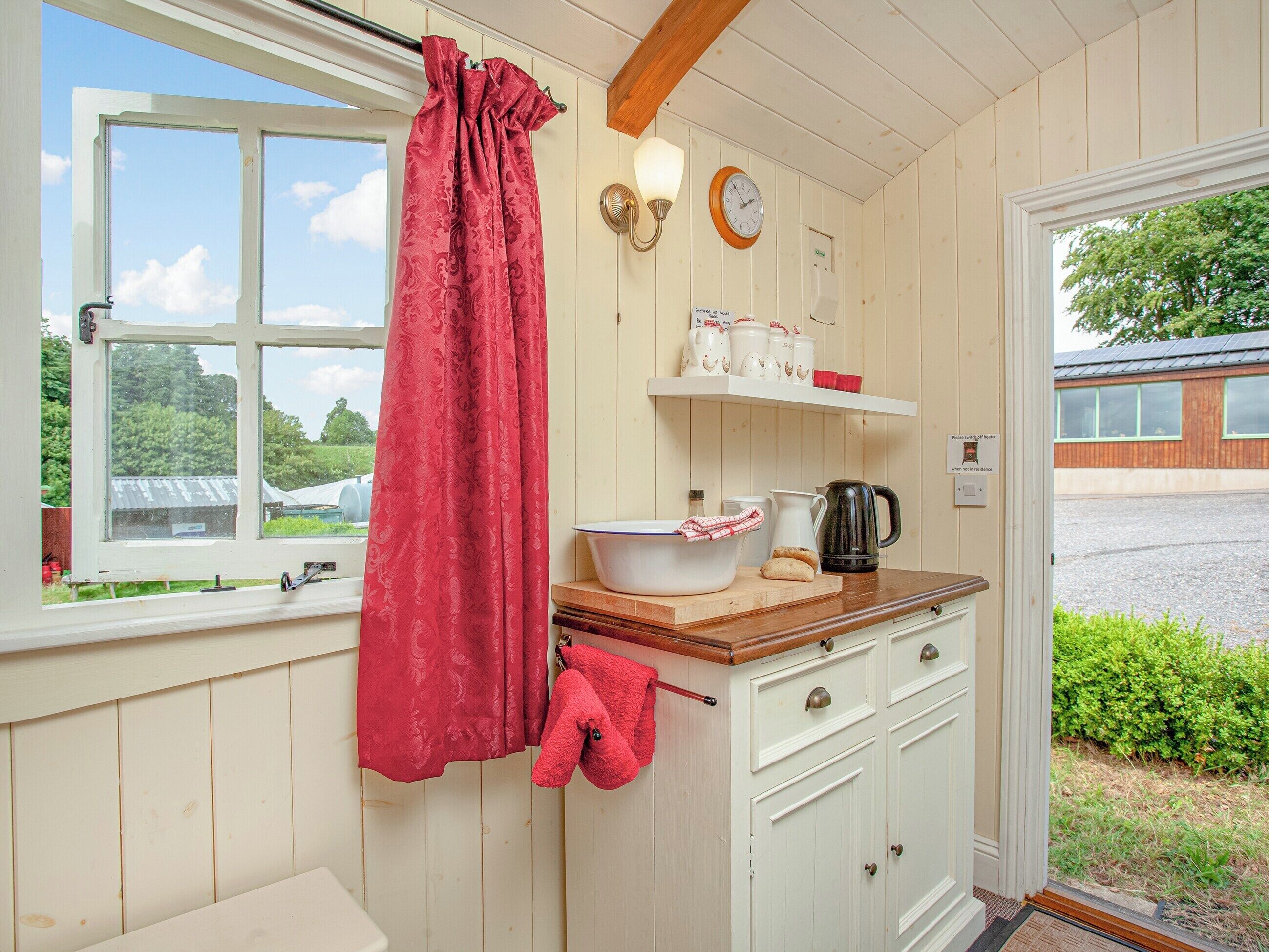 A kitchen with a window, a red curtain, a kettle, and a sink.