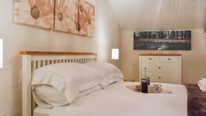 Ein ordentlich bezogenes Bett mit weißen Leinen, ein Nachttisch mit Tasse und Untertasse, ein Holzbeistelltisch, eine Kommode mit Spiegel und zwei Wandbilder.