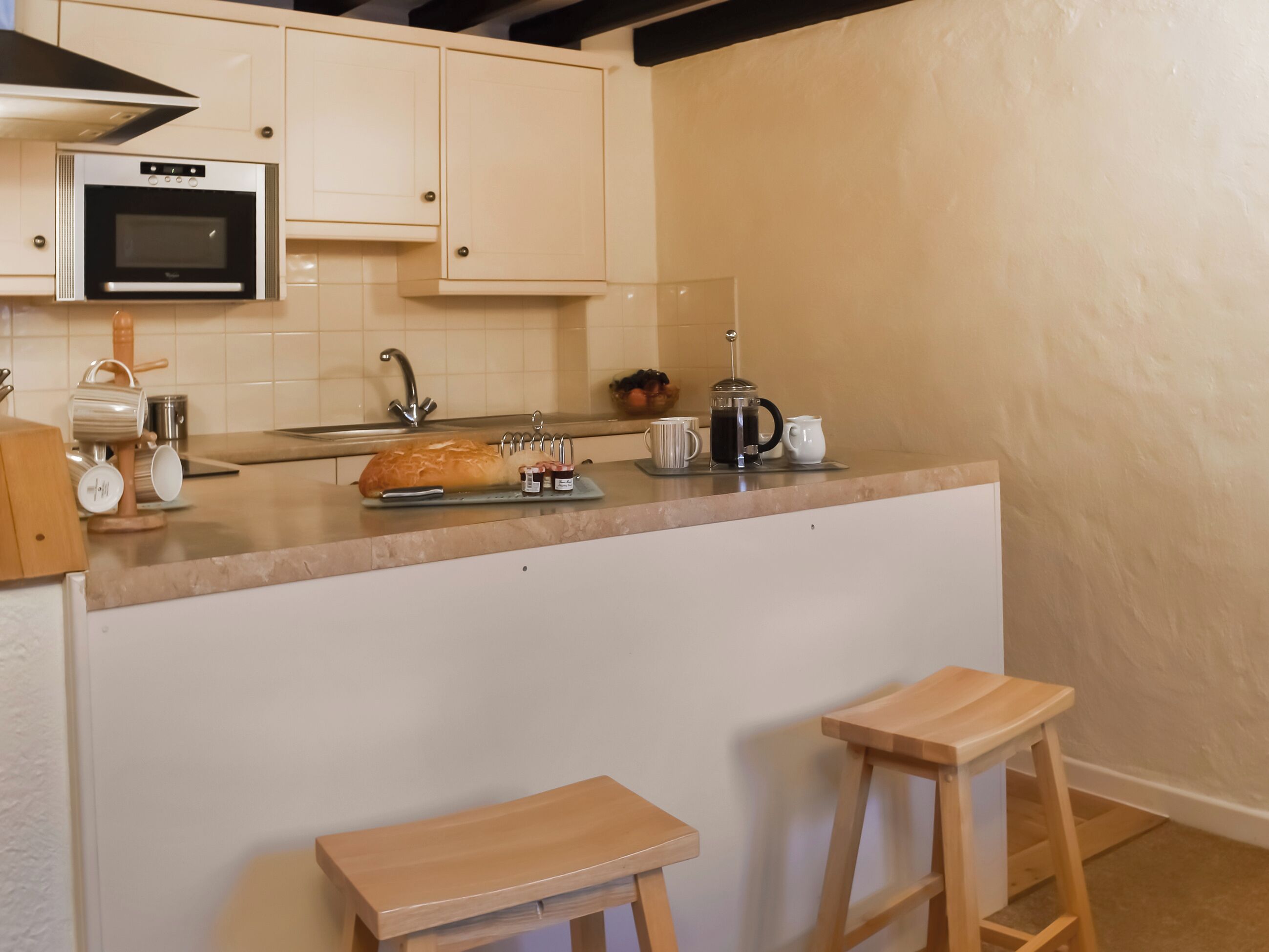 Een compacte keuken met een inbouwoven, een spoelbak en een koffiezetapparaat. Op het aanrecht staan twee houten krukken en een brood.