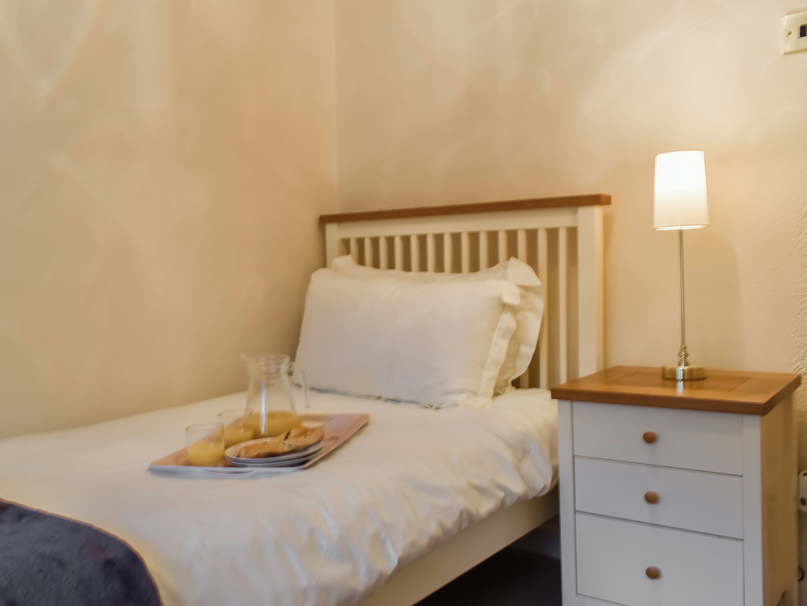 Ein ordentlich bezogenes Bett mit weißen Leinen, ein Nachttisch mit Lampe sowie ein Tablett mit Gebäck und Saft.