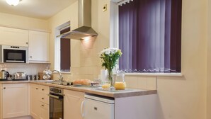Een kleine keuken met een magnetron, broodrooster, waterkoker en een spoelbak. Ook een koelkast, een fornuis en een raam met gordijnen.
