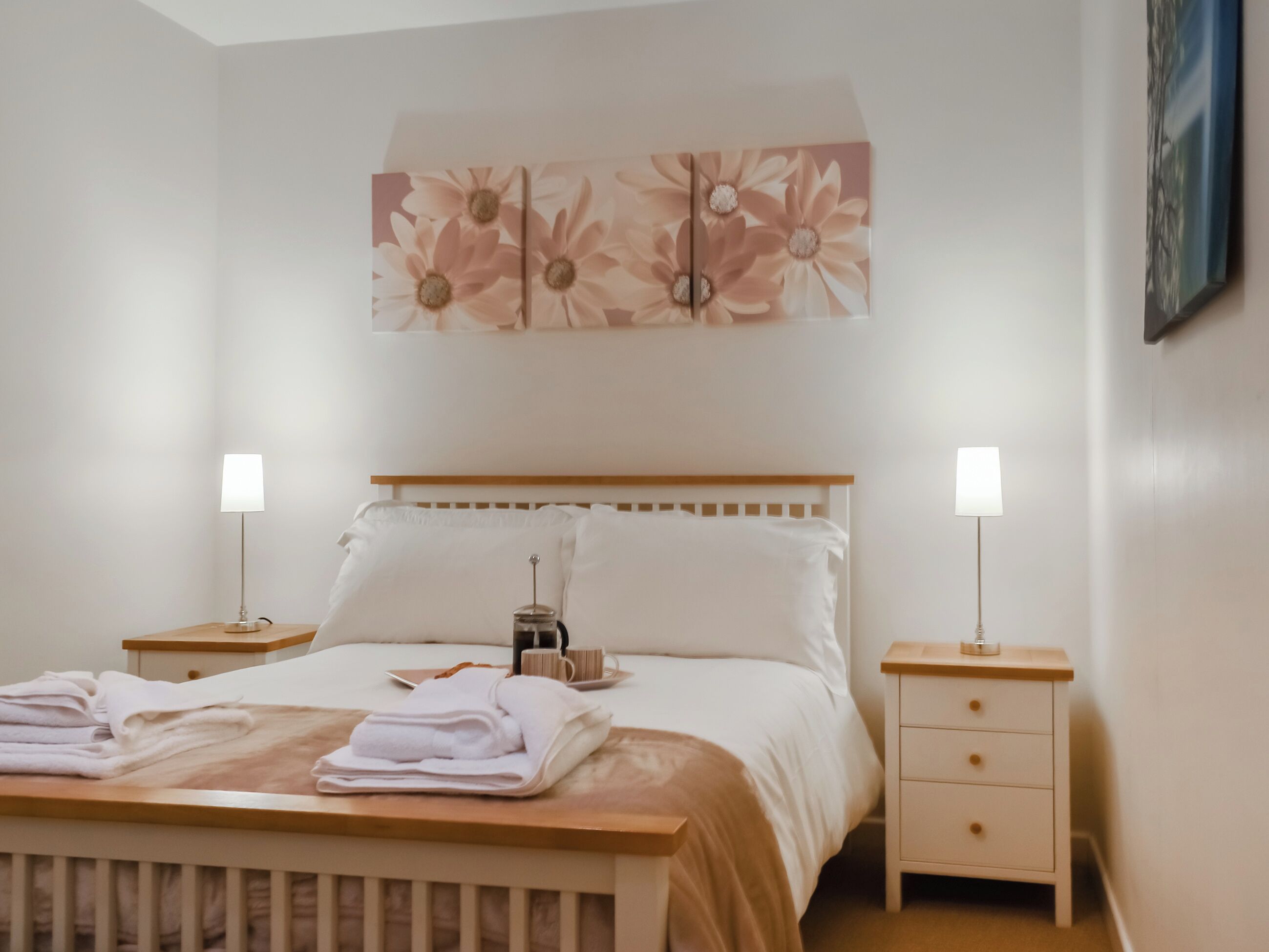 Un lit bien fait, avec une tête de lit en bois, deux tables de chevet avec des lampes et une œuvre d’art florale au-dessus du lit.