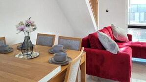 Ein moderner Essbereich mit Holztisch, grauen Stühlen und einer Vase mit Blumen. Im Hintergrund ist eine rote Couch mit Kissen zu sehen.