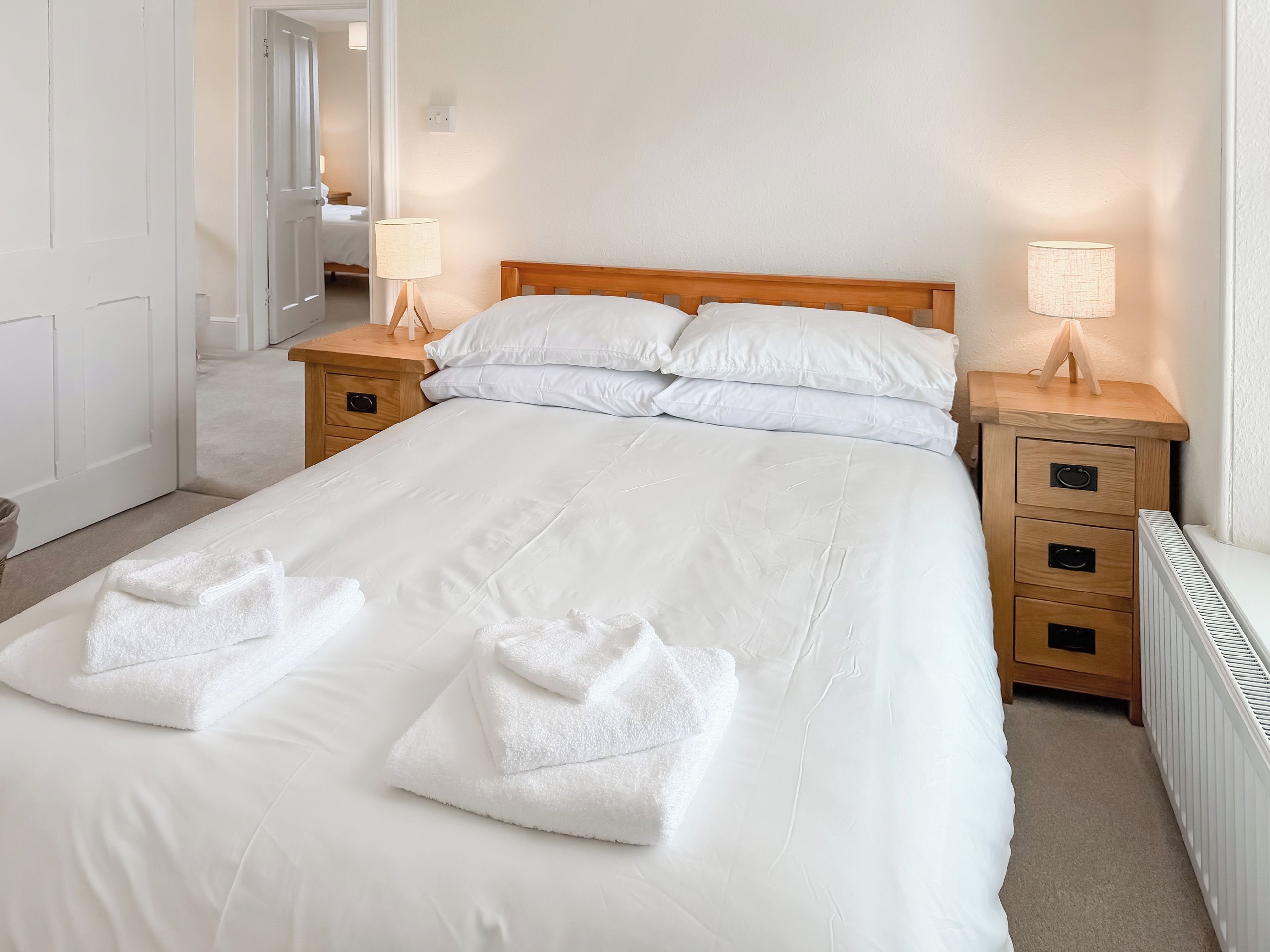 A neatly made bed with white linens and towels, two bedside tables with lamps, and a white radiator.