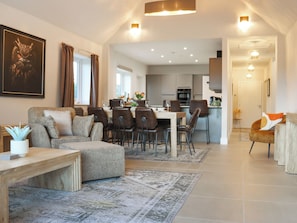 Ein geräumiger Wohnbereich mit einer Couch, einem Esstisch und Stühlen, eine Küche im Hintergrund und ein Flur, der zu einem weiteren Raum führt.