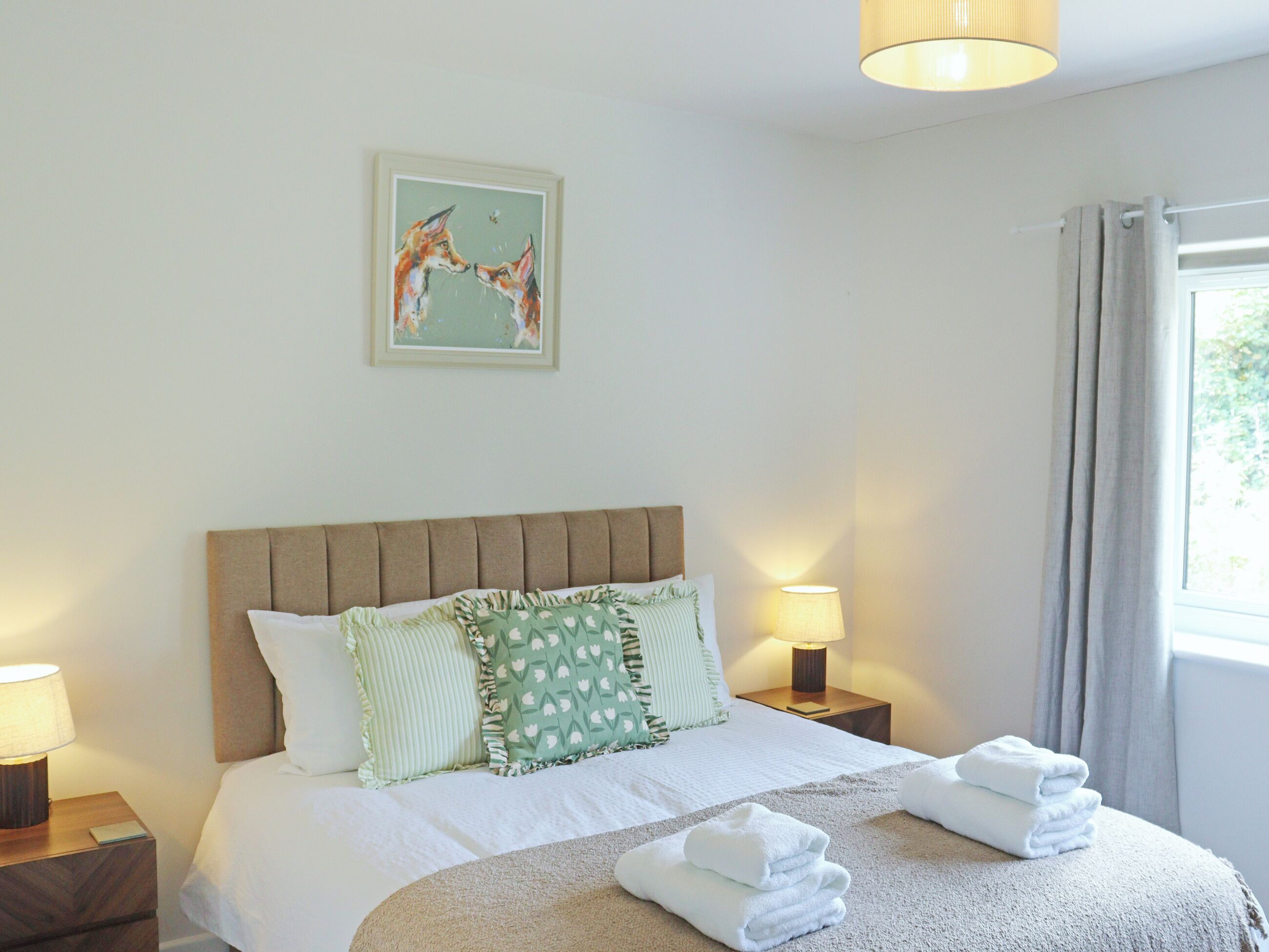 A hotel room with a bed, pillows, a nightstand with a lamp, a painting on the wall, and a window with curtains.
