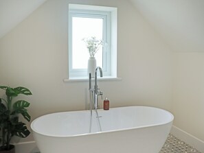Eine weiße, freistehende Badewanne mit einem Fenster darüber, eine Topfpflanze links und eine Seifenflasche am Rand.