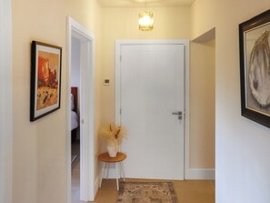 Un couloir avec une porte blanche, un tableau encadré sur le mur de gauche, un vase contenant des plantes séchées sur une petite table, et un tapis à motifs sur le sol.