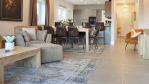 A spacious living area with a sofa, dining table, and chairs, a kitchen in the background, and a hallway leading to another room.