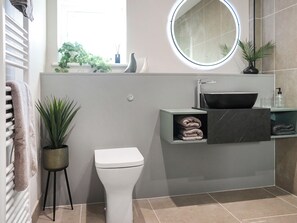 A modern bathroom with a toilet, a sink with a vessel basin, a round mirror, and a towel rack. - Spacious, beautifully finished home with private hot tub and countryside views. (Haverfordwest)