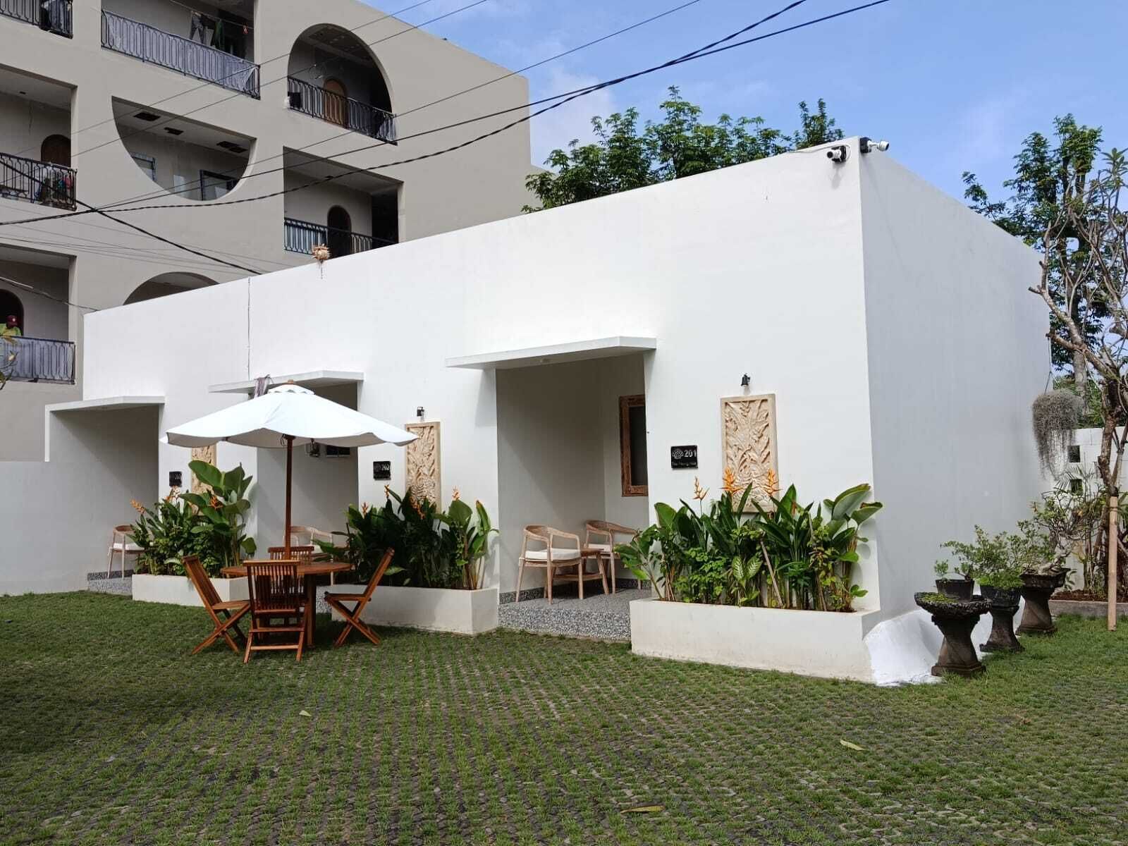 A modern building with a flat roof, white exterior walls, and a paved courtyard. There are outdoor seating areas with wooden chairs and tables, and several potted plants.