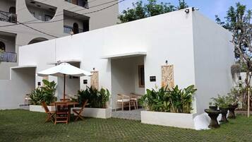 A modern building with a flat roof, white exterior walls, and a paved courtyard. There are outdoor seating areas with wooden chairs and tables, and several potted plants.