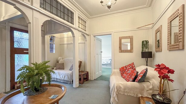 A spacious living room with a high ceiling, a dining area with a wooden table and chairs, a cozy sofa with red and white cushions, and a potted plant.
