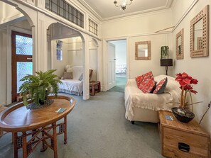 A spacious living room with a high ceiling, a dining area with a wooden table and chairs, a cozy sofa with red and white cushions, and a potted plant.