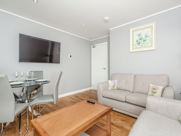 A modern living room with a sofa, a dining table with chairs, and a flat-screen TV mounted on the wall. - Inverness Highland Apartment (Inverness)