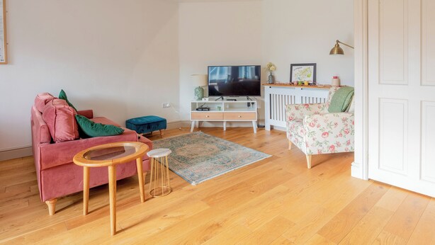 Una sala de estar con un sofá rosa, un reposapiés azul, una mesa de centro de madera, un televisor, un armario blanco y un sillón con estampado floral.