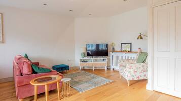 A living room with a pink sofa, a blue ottoman, a wooden coffee table, a TV, a white cabinet, and a floral armchair.