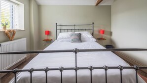 A bedroom with a bed, two bedside tables, a window with blinds, a radiator, and a ceiling light fixture.