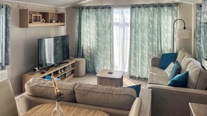 A modern living room with a flat-screen TV, a wooden entertainment unit, a sofa set, and a dining table with a vase. - 3 bedroom accommodation in New Quay (New Quay)