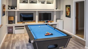 Een pooltafel met een blauwe viltbekleding, omringd door planken met diverse spullen, een televisie en een deur naar een andere kamer.