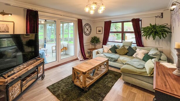 A living room with a television, a wooden coffee table, a sofa with cushions, and a large window with curtains. - 1 bedroom accommodation in Abergele (Abergele)