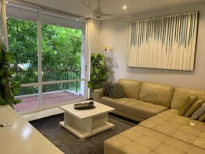 Ein modernes Wohnzimmer mit einem beige gefärbten Sofa, einem weißen Couchtisch und einem großen Fenster mit Blick auf Grünfläche.