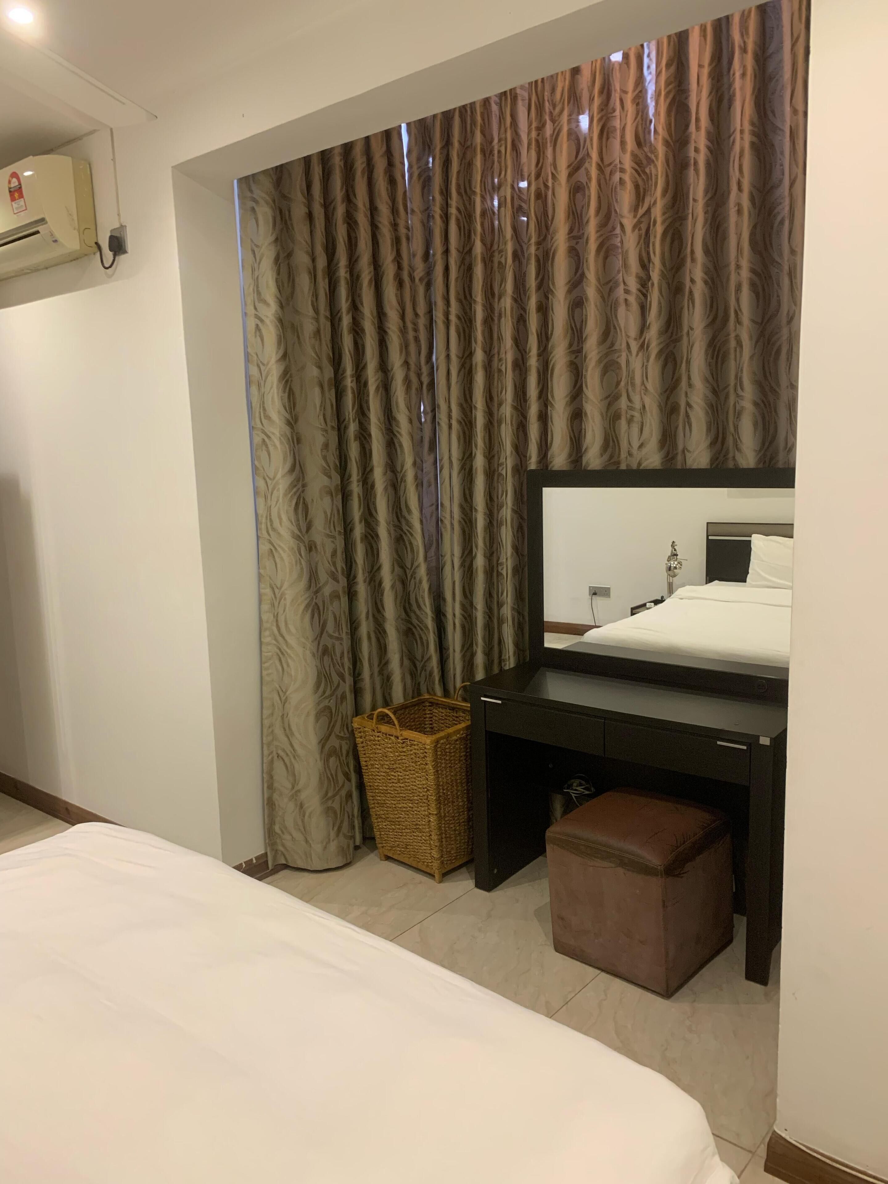 A hotel room with a bed, a desk with a mirror, a small wicker basket, and a brown ottoman.