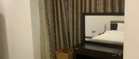 A hotel room with a bed, a desk with a mirror, a small wicker basket, and a brown ottoman.