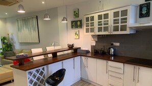 Cocina moderna con armarios blancos, encimera de madera e iluminación colgante. Hay un taburete de bar y una mesa de comedor con sillas.