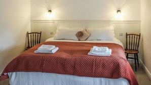 Ein ordentlich bezogenes Bett mit einer roten Steude, zwei weißen Kissen und einem kissenförmigen Rosenmotiv. Zwei Holzstühle stehen daneben.